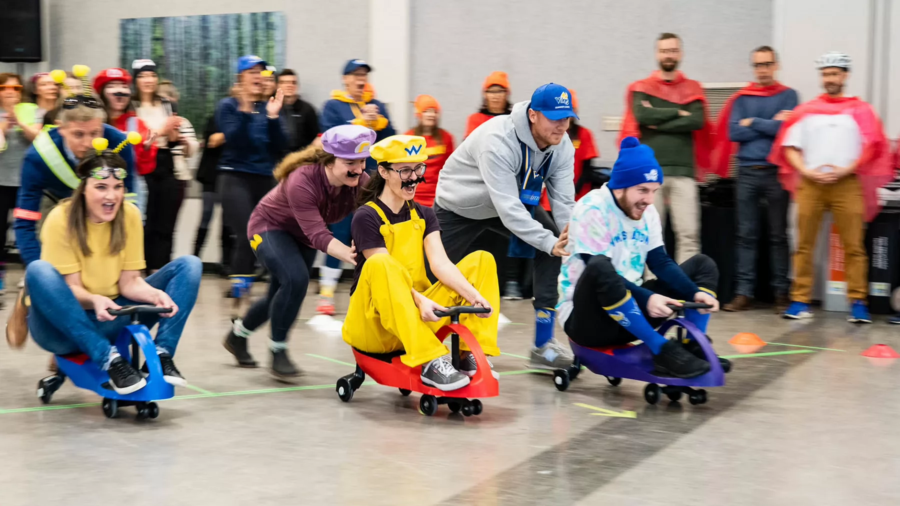 A crowd of people cheer on teams of plasma car racers wearing costumes.