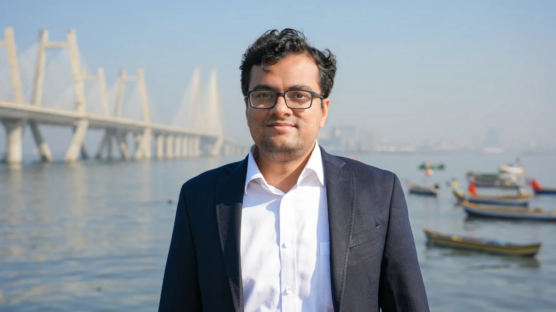 A man in a suit standing in front of a body of water with a large bridge behind him.
