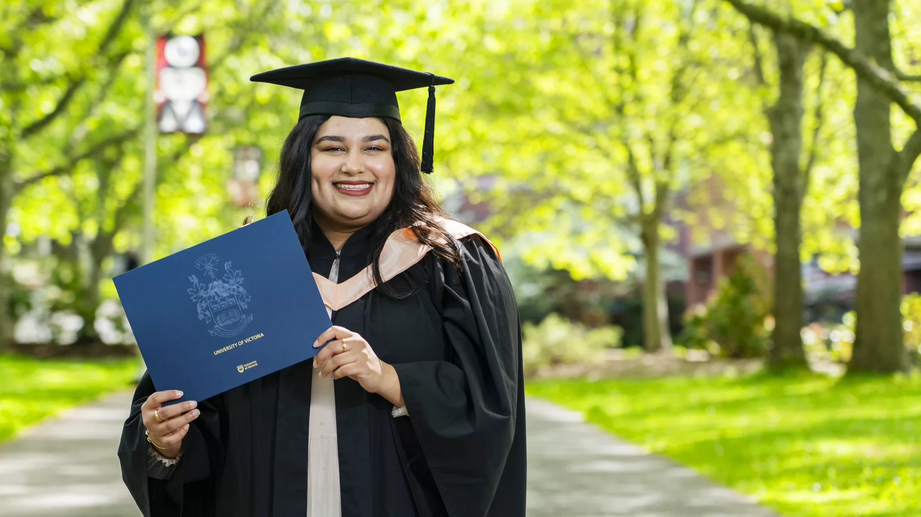 A person dressed in academic regalia (cap and gown) smiles, holding a degree cover, standing on a path surrounded by trees.