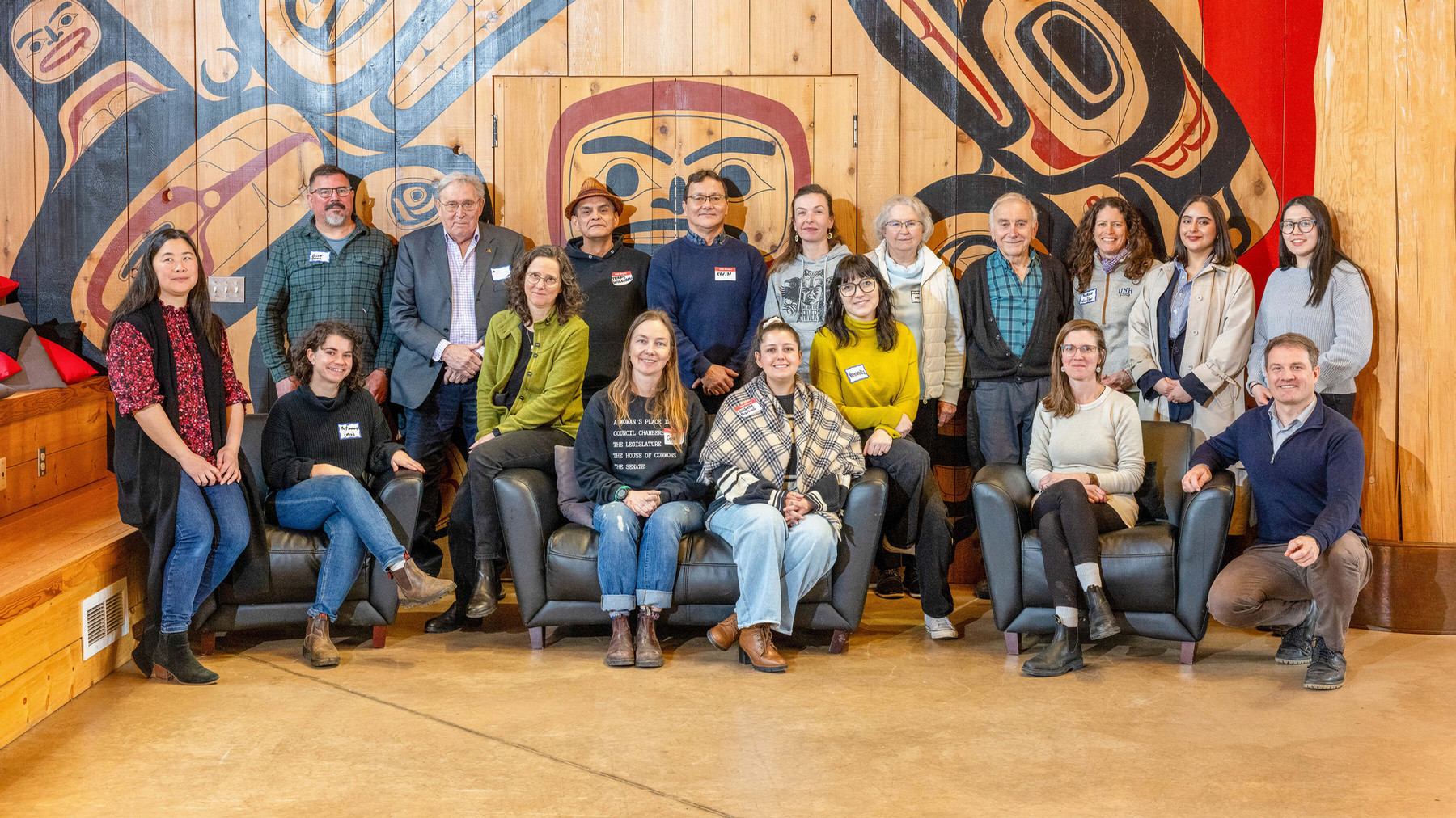 Eighteen individuals of various ages and backgrounds pose for a group photo beneath a mural painted in the traditional Haisla style.