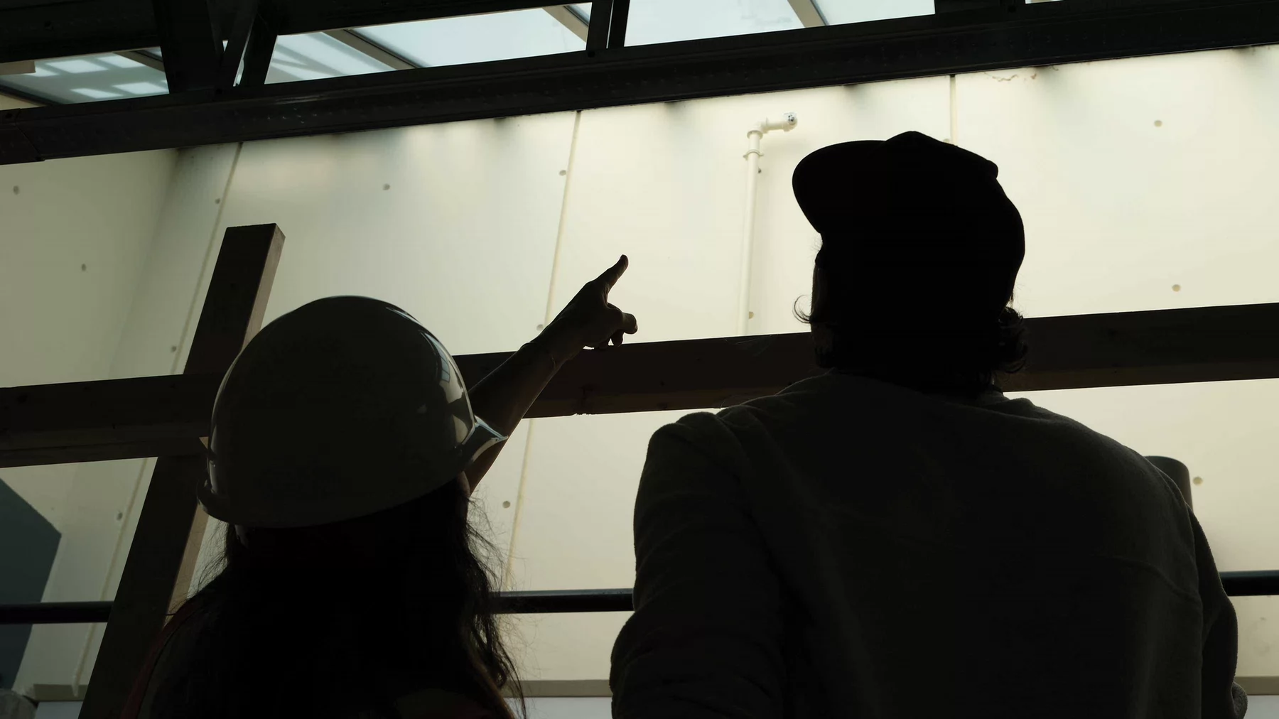 Two people in silhouette, one wearing a hard hat, stand indoors beneath a glass ceiling and looking up as one points toward overhead structures.