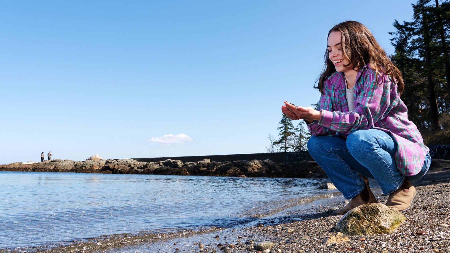 UVic student Quinn Bitz crouches on a rocky beach, holding pebbles in her hand