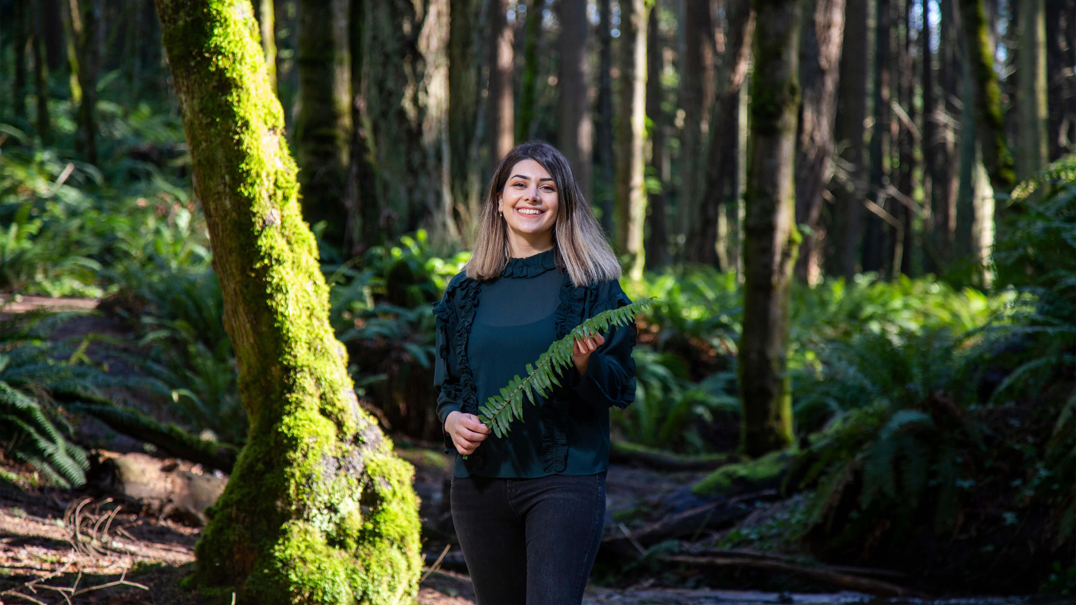 A woman stands in a forest holding a fern and smiling