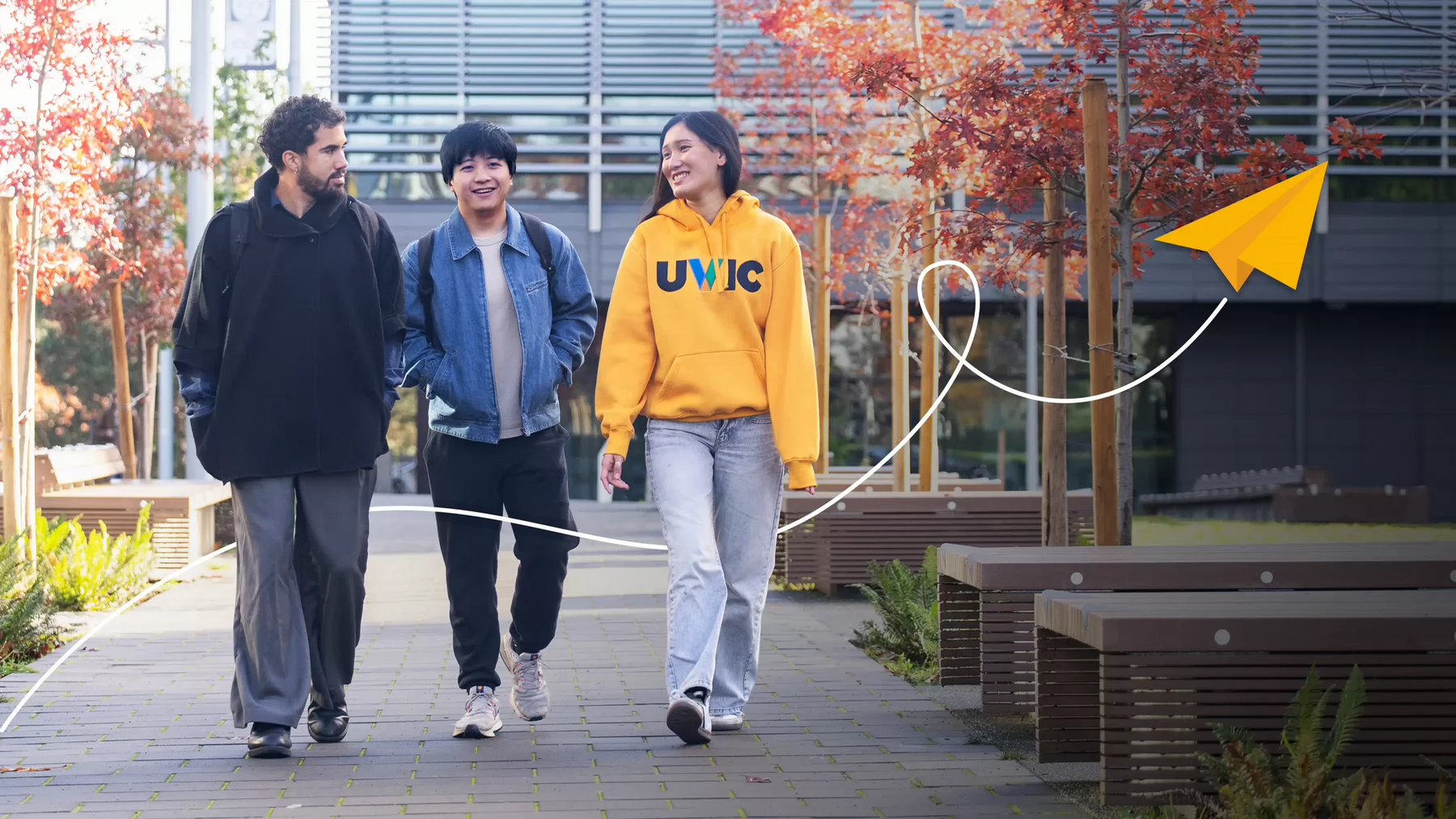 Three students walk outside the new residence building in autumn. An illustrated paper plane flies by. 