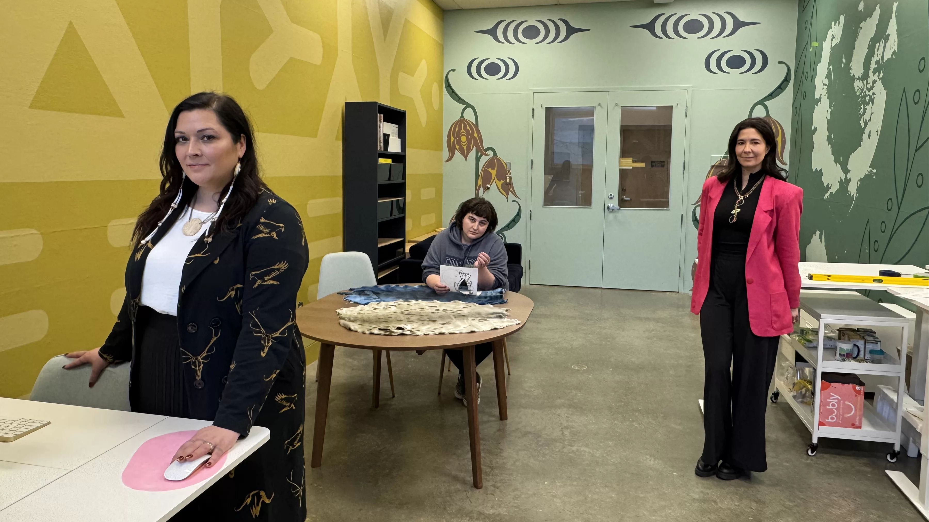 Heather Igloliorte stands in the Taqsiqtuut Research-Creation Lab. Another person sits at a table behind her and another person stands by a shelf.