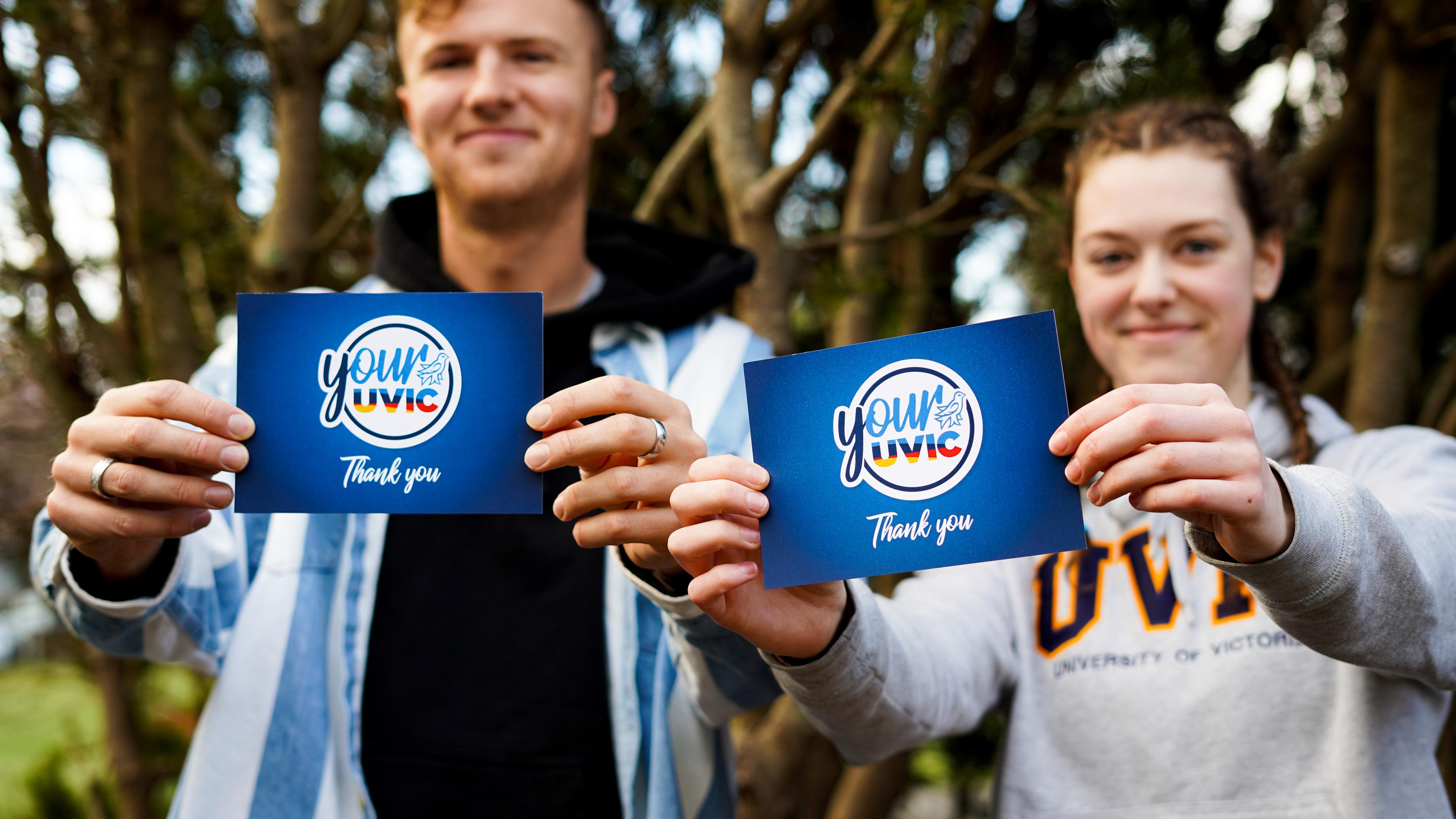 Two students smile holding your uvic thank you cards in front of them