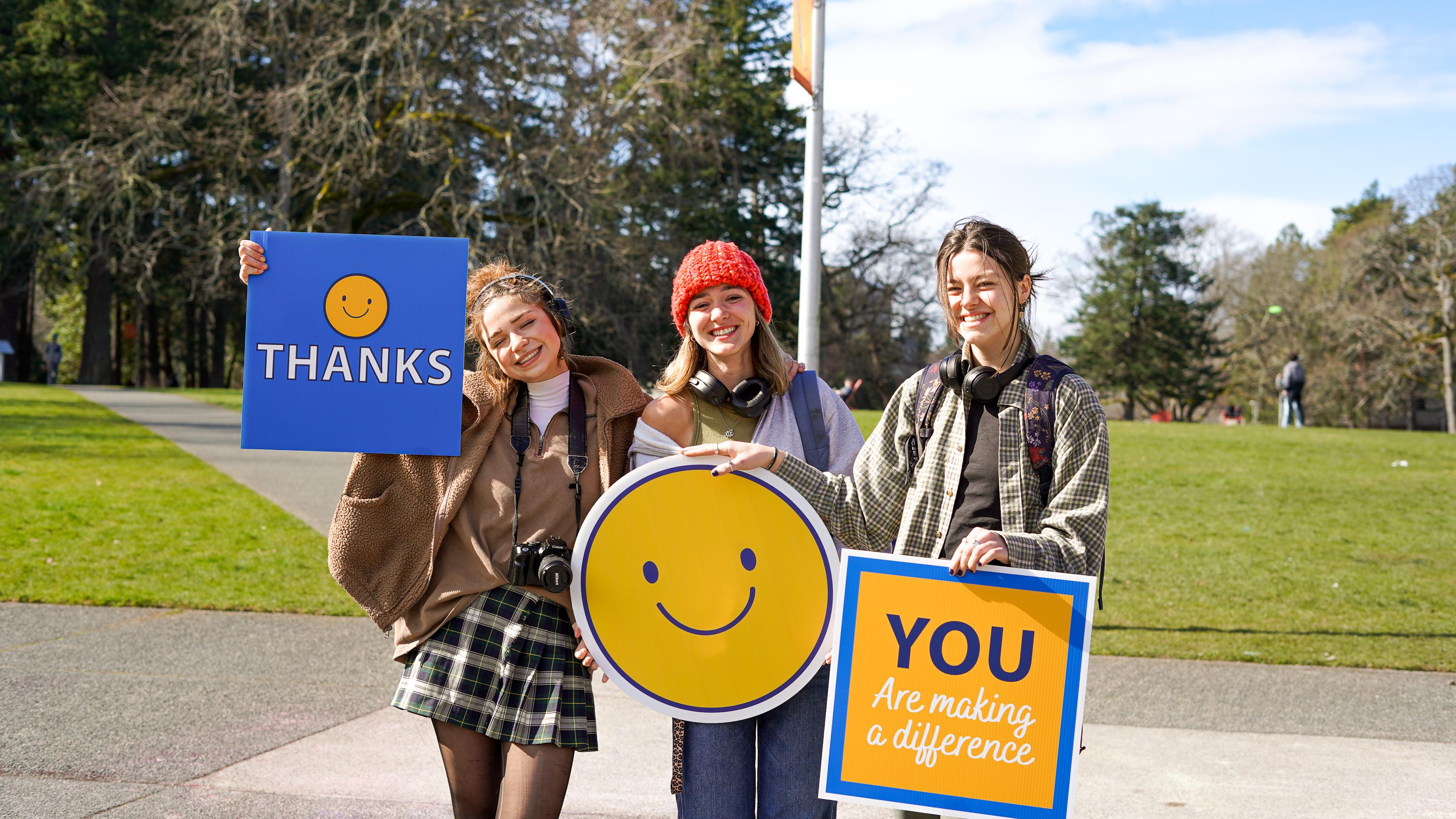 Three female students stand together smiling and holding signs, from left to right the signs are "Thanks", a giant yellow smiley, "you are making a difference" 