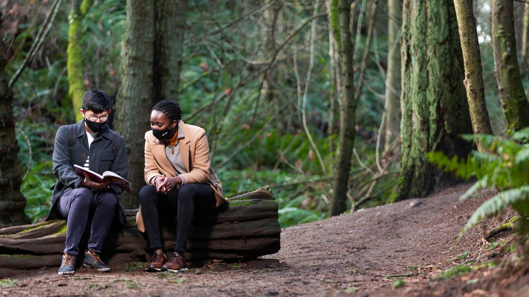 Two masked students sit on a log in Mystic Vale looking at a book