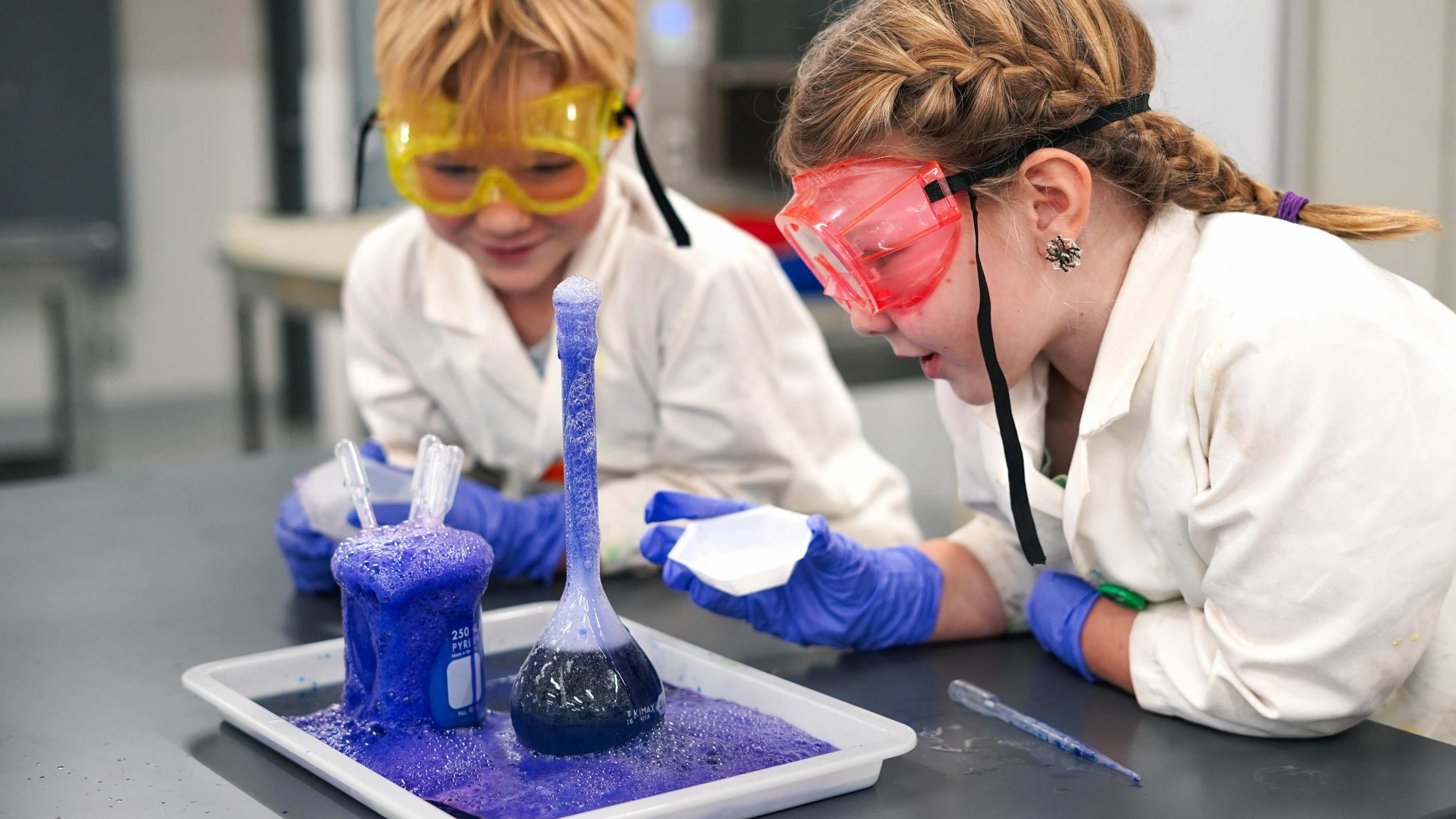 Two six year-old kids wearing lab coats and eyewear lean over a science beaker of purple bubbling liquid.