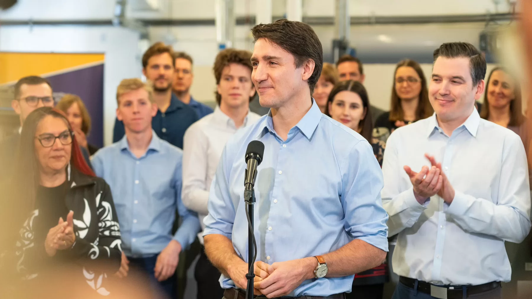 Prime Minister Justin Trudeau stands at a microphone stand smiling with people standing behind. 