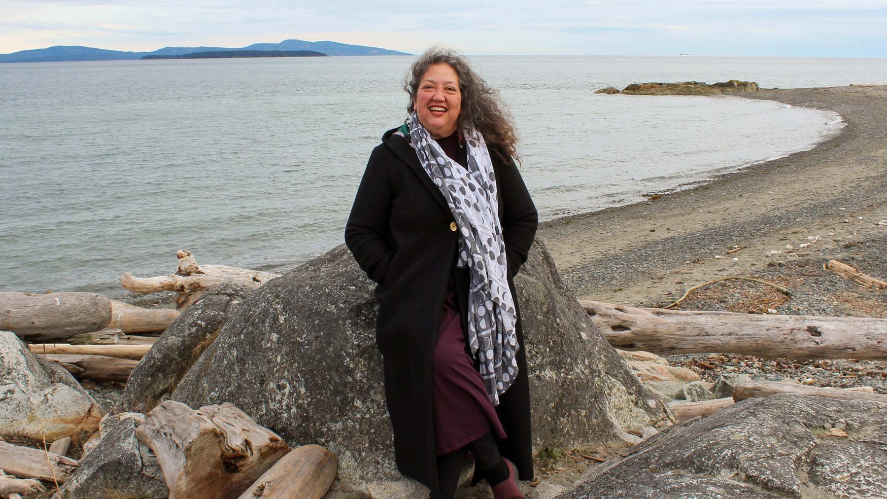 Lisa Kealohapaʻaokahaleole Hall stands on the beach and smiles at camera.