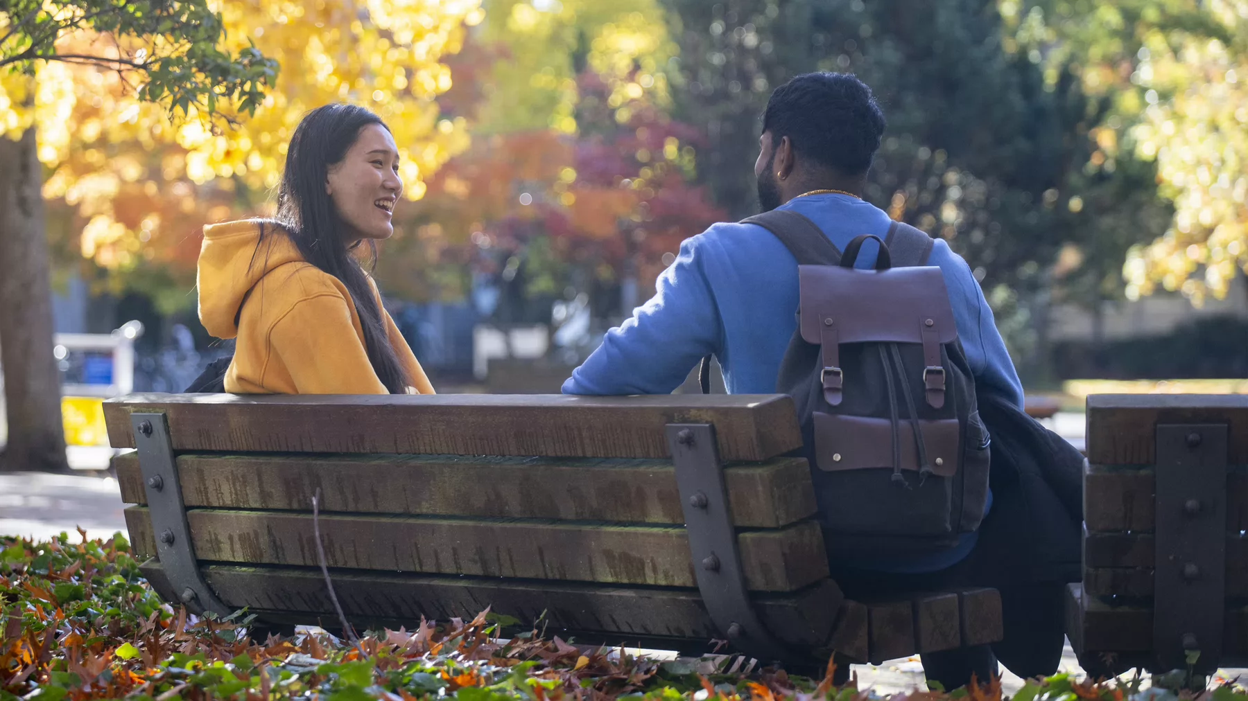 Two students sit on a bench surrounded by trees with fall coloured leaves.