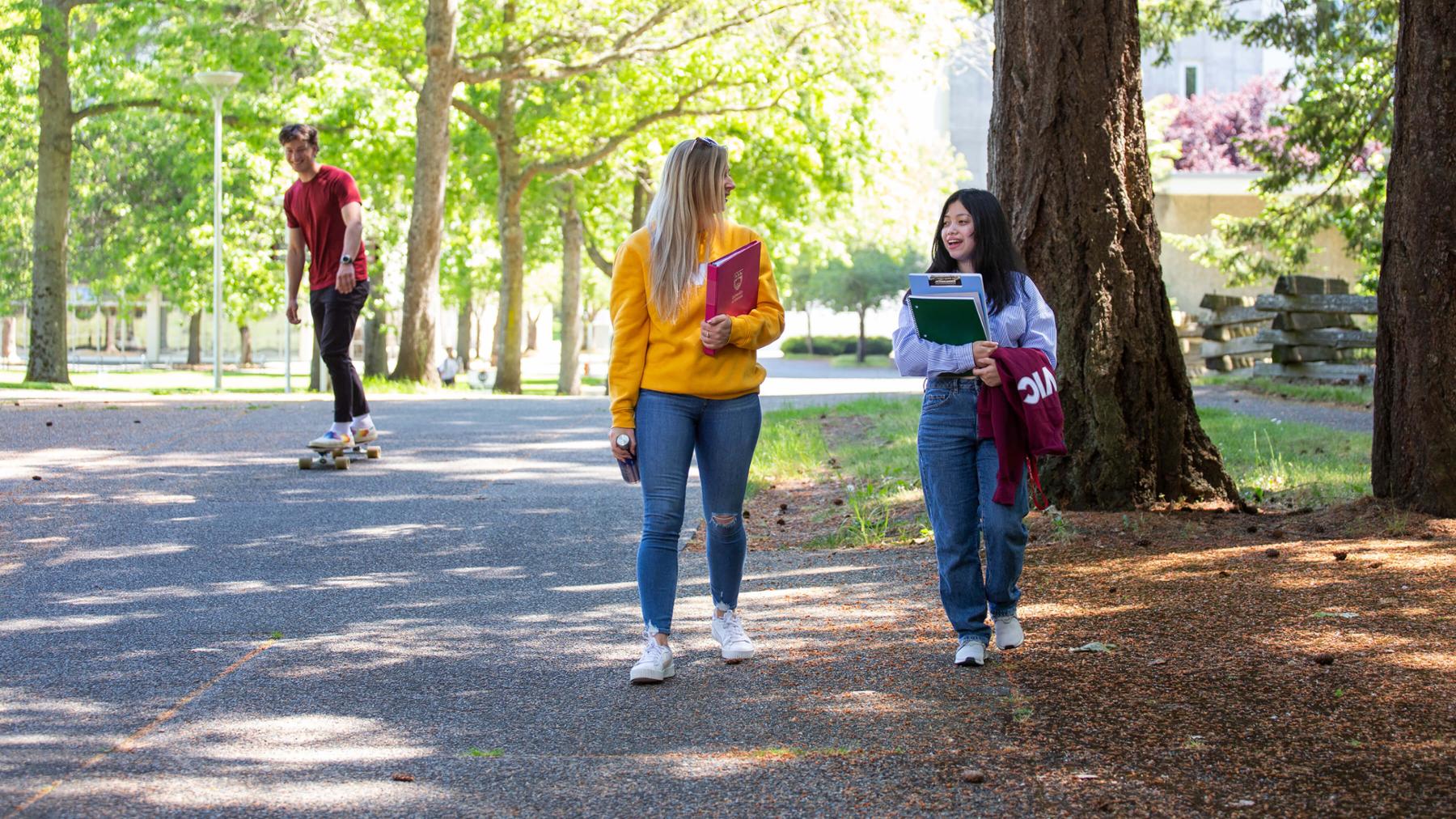 Two students walk along a pathway on campus, while another student rides a skateboard in the background.