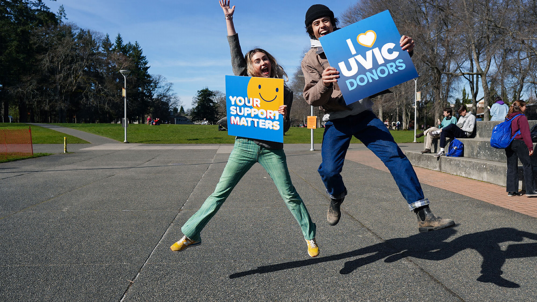 Two students smiling and jumping holding signs that read "your support matters" and "I heart UVic donors"