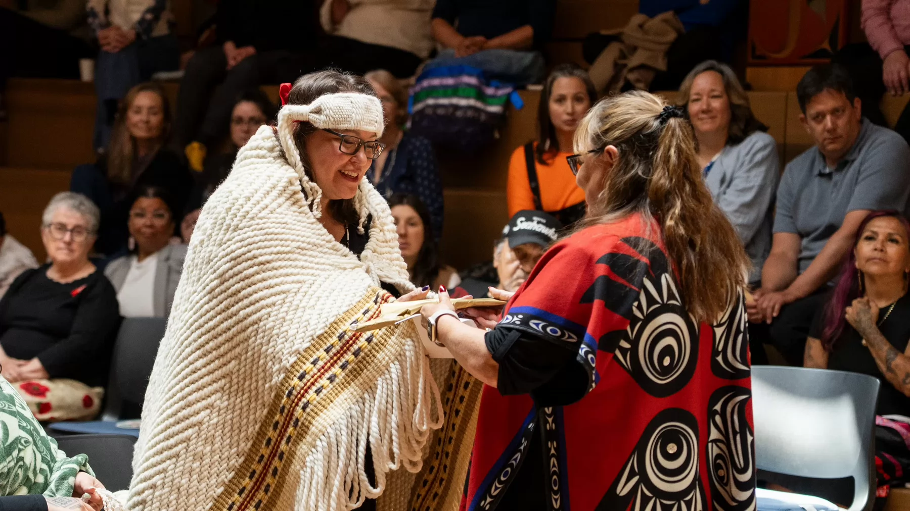 Indigenous woman wearing a woven blanket accepts a beaded feather gift from another woman wearing a red and black Indigenous form line shawl.