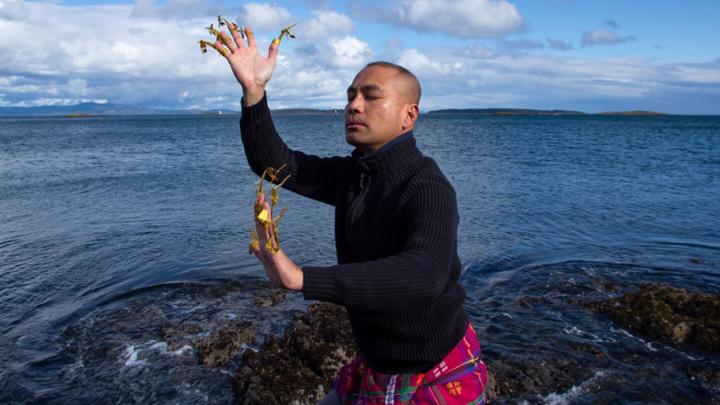 Male dancer with golden finger adornments mid-pose on a rock by the ocean