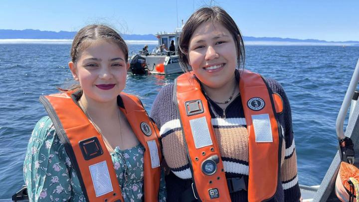 Two young Inuit women in lifejackets stand on a boat with the sea in the background.