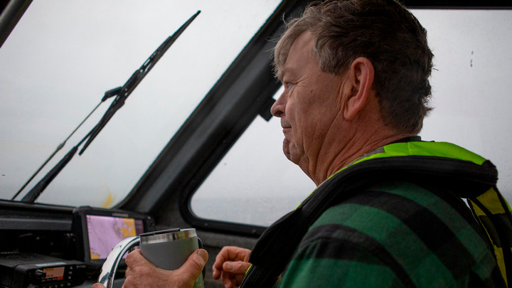 A side-profile portrait of Roger Dunlap at the wheel of his boat, coffee mug in hand