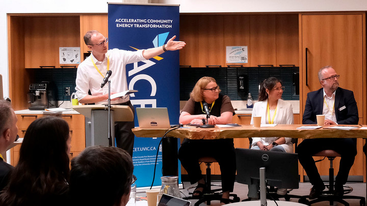 ACET ED Curran Crawford stands at a podium and gestures toward a panel of three speakers seated at a live-edge wooden table during a community energy conference. Microphones, a laptop, and coffee cups are on the table, and a banner reading "Accelerating Community Energy Transformation" stands behind the speakers as attendees listen from the audience.