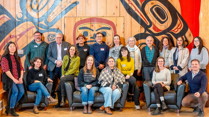 Eighteen individuals of various ages and backgrounds pose for a group photo beneath a mural painted in the traditional Haisla style.