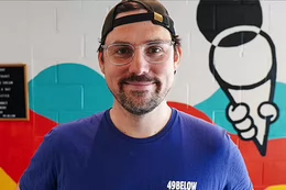 Man wearing glasses, backwards baseball and T-shirt that says 49 Below Ice Cream smiling while standing in front of colourful mural of an ice cream cone.  