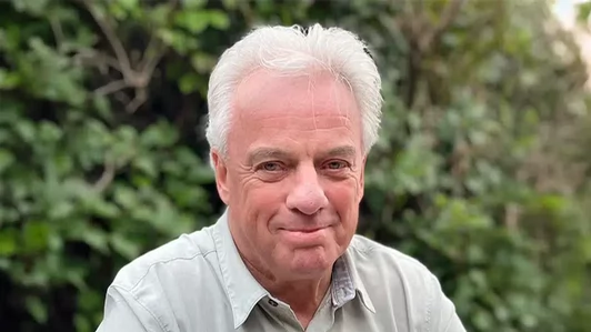 A man with white hair wearing a collared shirt and smiling.