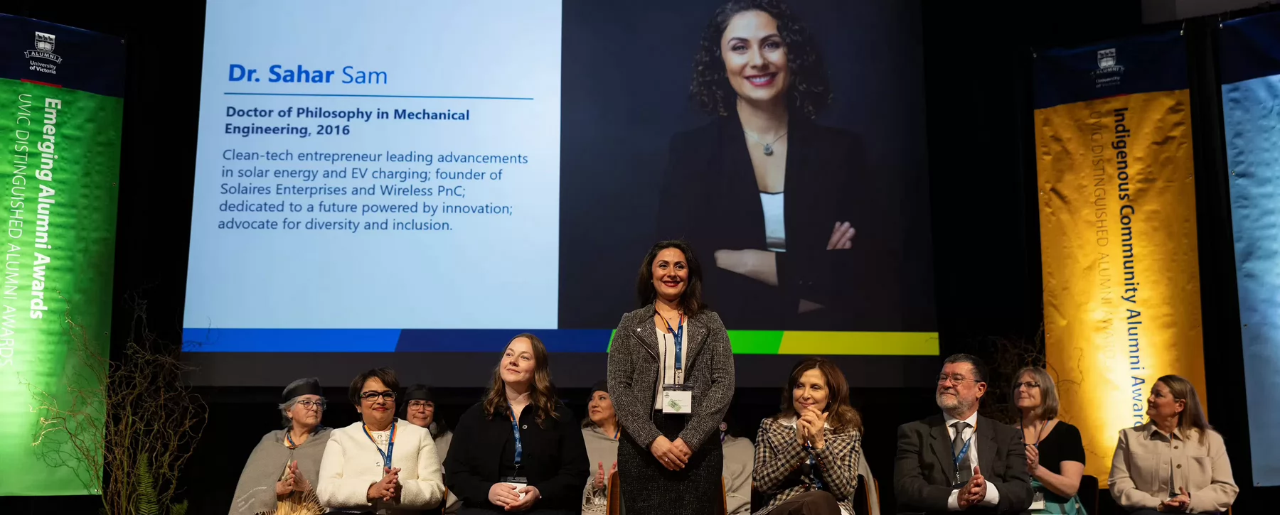 A woman standing and being honoured on a stage for an award with fellow award recipients who are sitting down and an overhead screen that says Dr. Sahar Sam, Emerging Alumni Award.