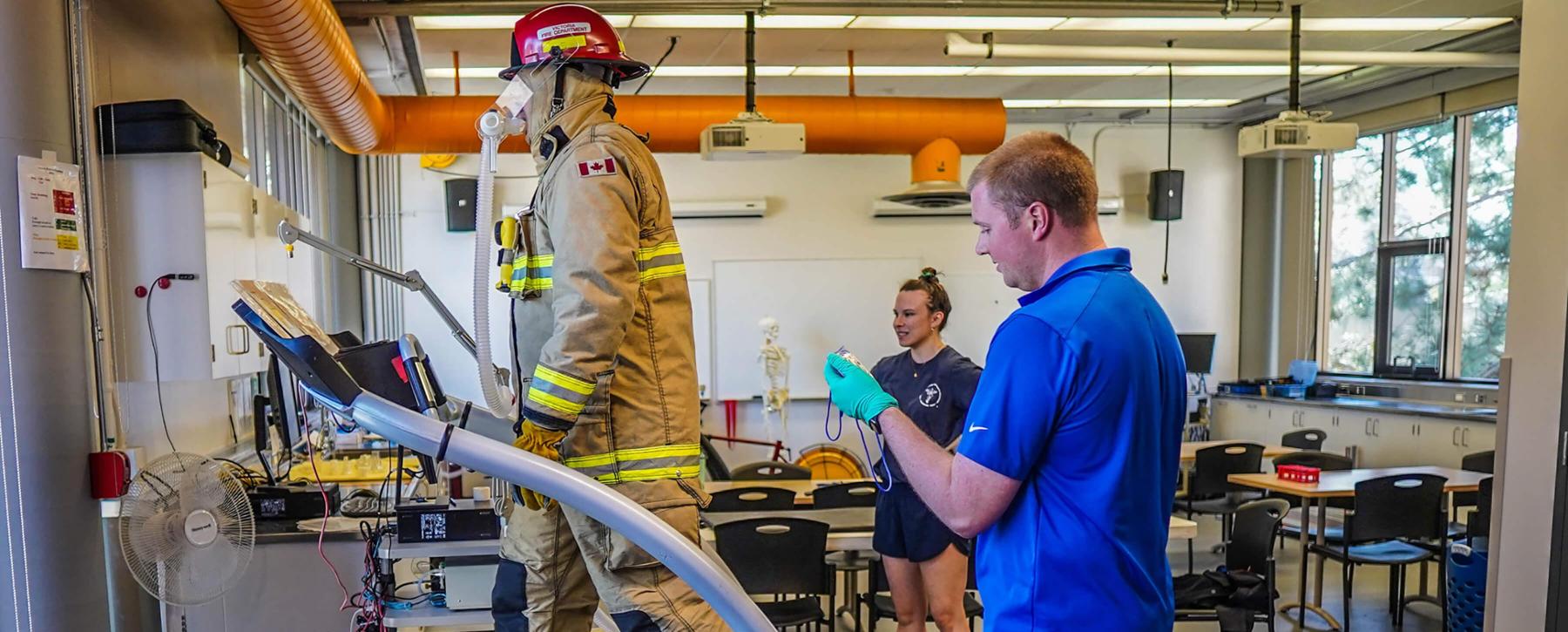 Man and woman monitor a firefighter in full gear walking on a treadmill. 
