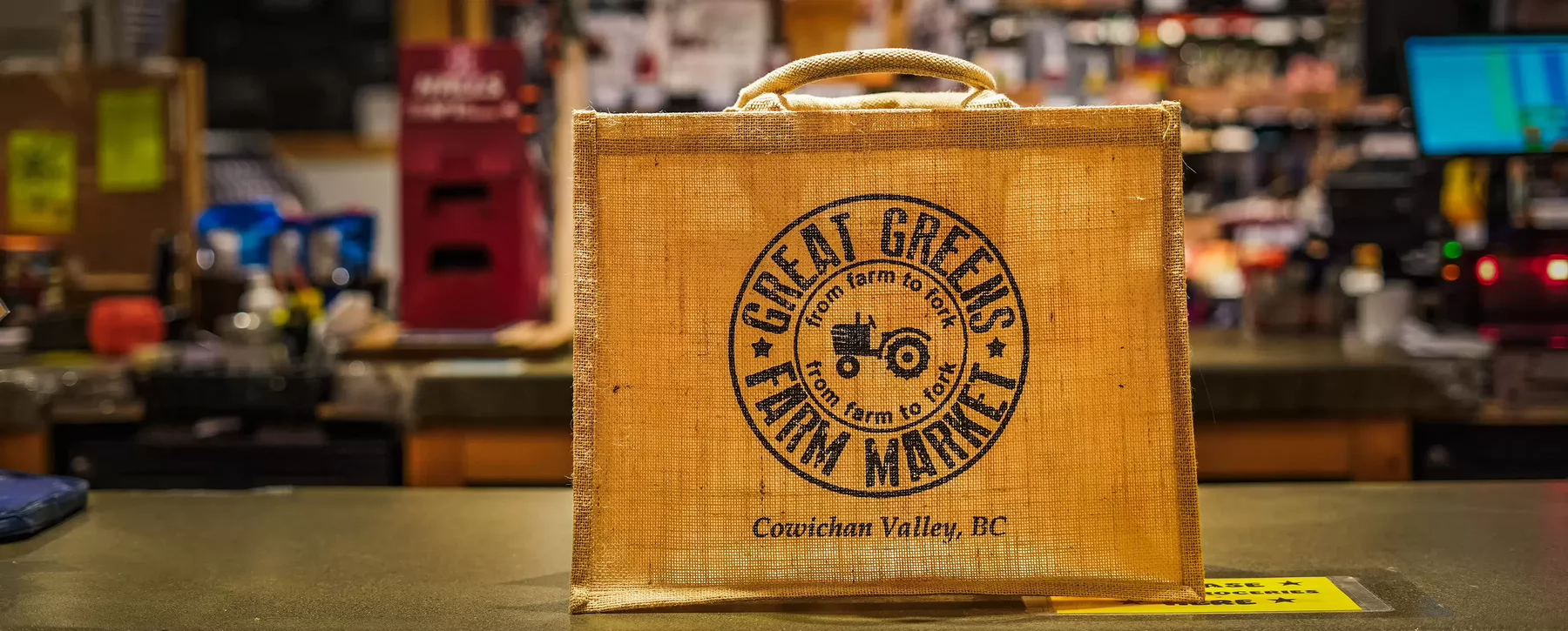 A burlap shopping bag that says Great Greens Farm Market sitting atop a counter in a grocery store.