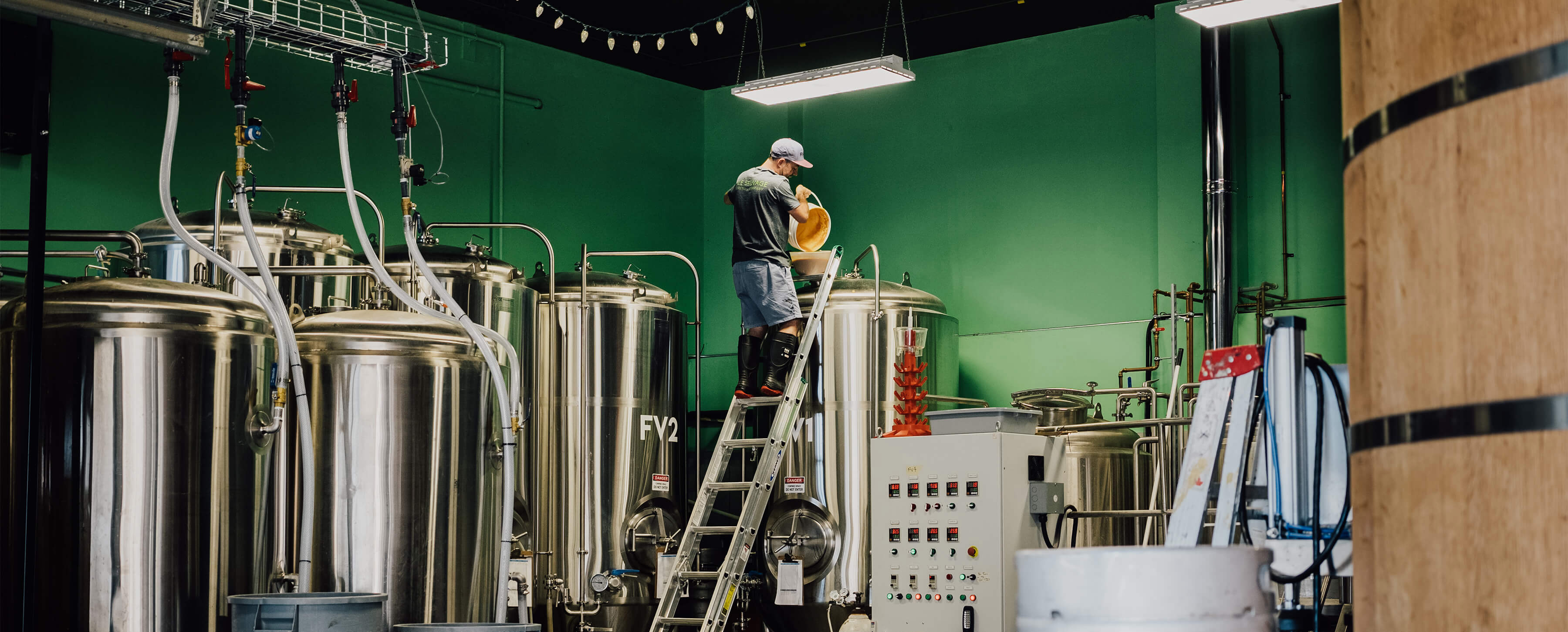 Man in baseball hat, shorts and rubber boots stands on a ladder pouring contents of a bucket into a tall beer tank.