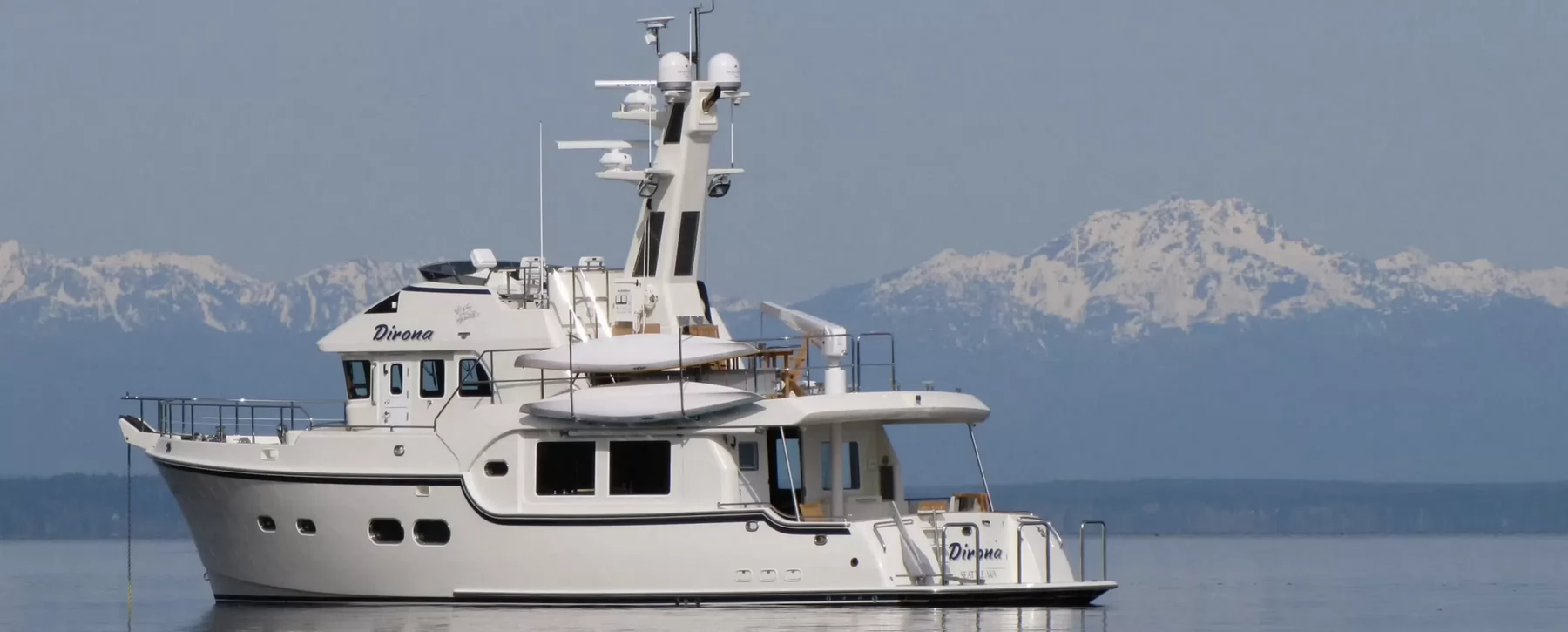 A yacht named Dirona floating on a calm ocean with snowcapped mountains in the background.