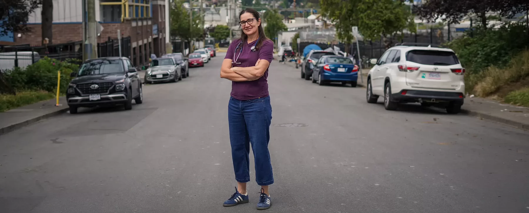 A woman wearing glasses and a stethoscope around her neck standing with her arms crossed in the middle of a road.