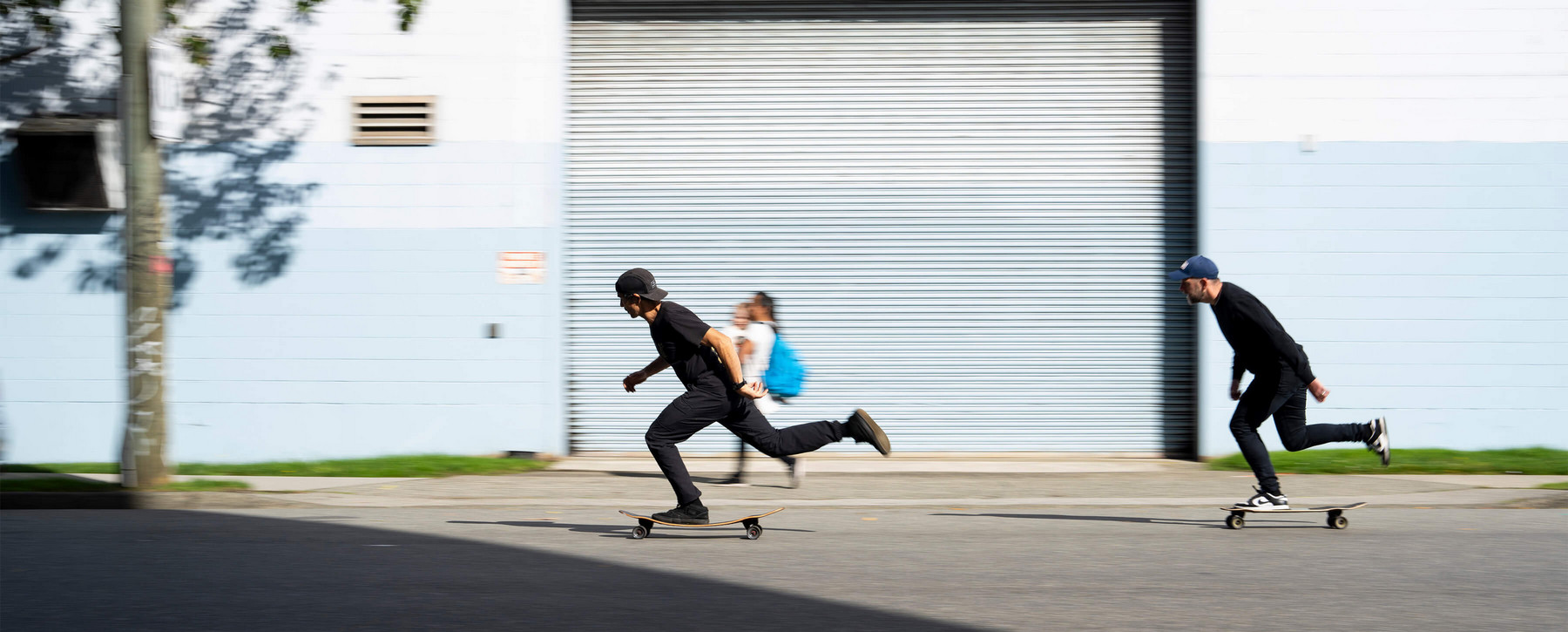 Two people skateboard past an industrial building. 