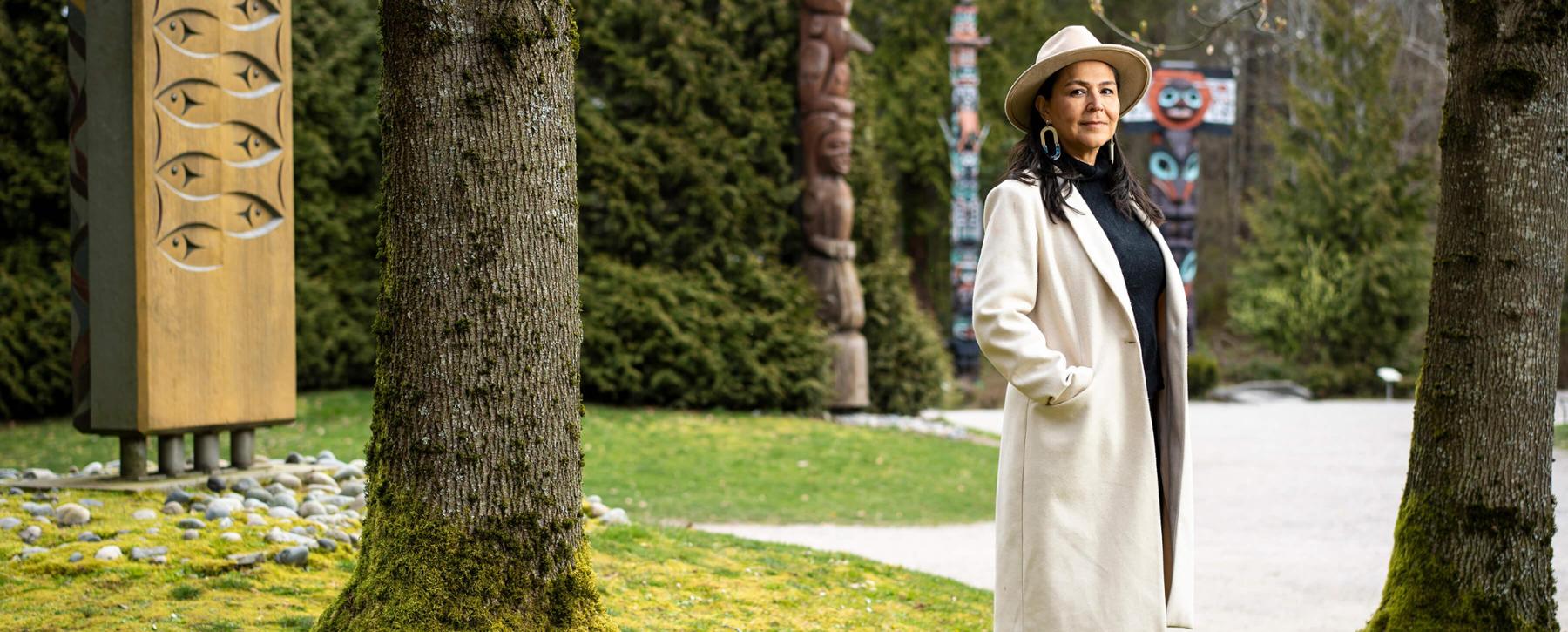 Woman wearing brimmed hat and long coat standing in front of totem poles. 