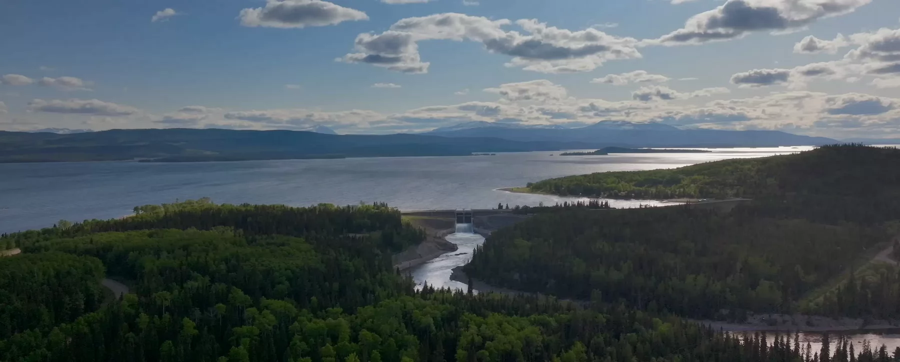 An aerial shot of a river winding through a forested area to a larger body of water separated by a dam.