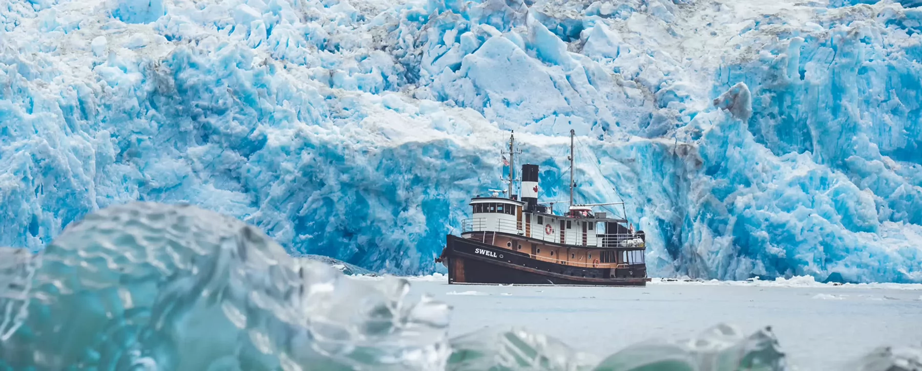 An old ship called Swell floating on the water in front of a glacier. 