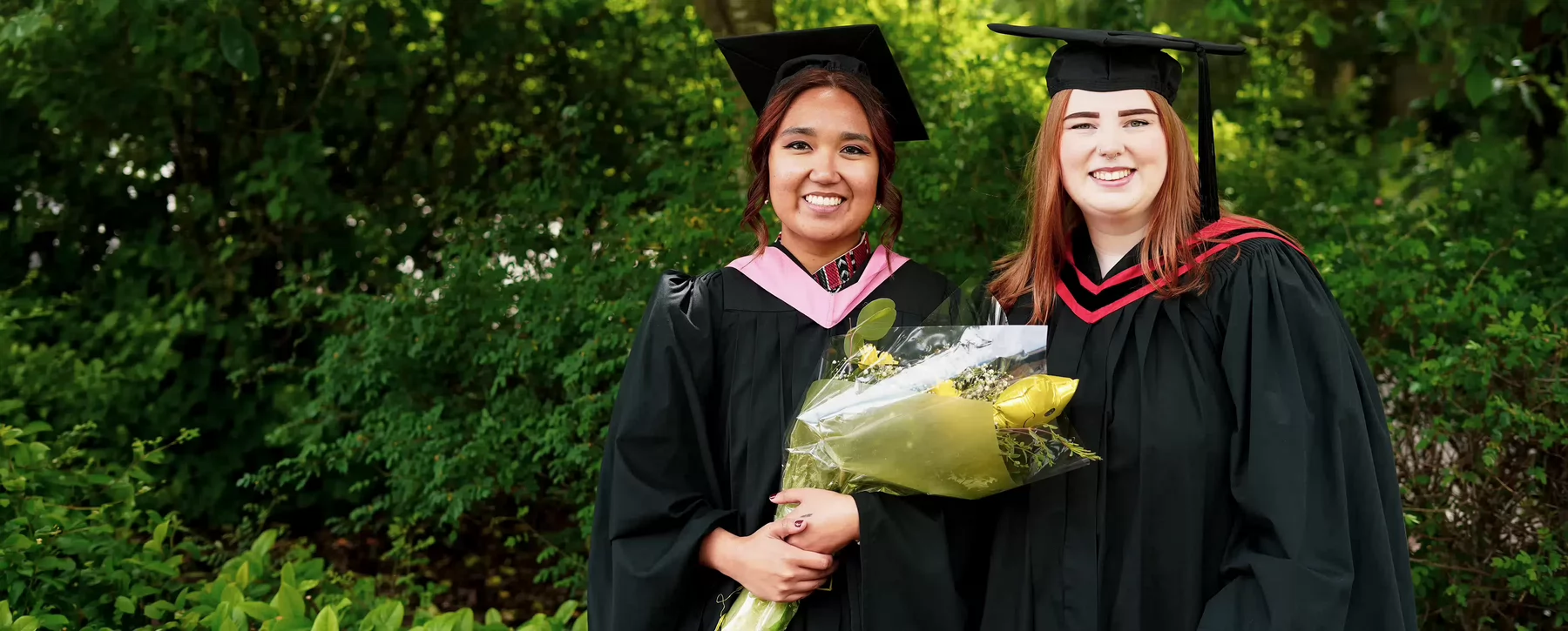 Two university graduates wearing caps and gowns smiling while standing in front of shrubbery. .