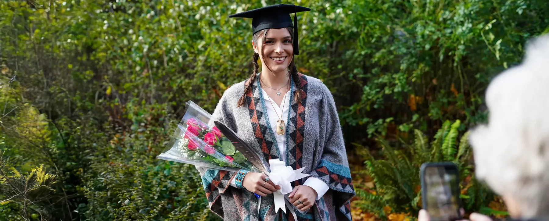 Woman wrapped in a Indigenous blanket, wearing graduation cap and smiling for camera while holding a feather and flowers.  
