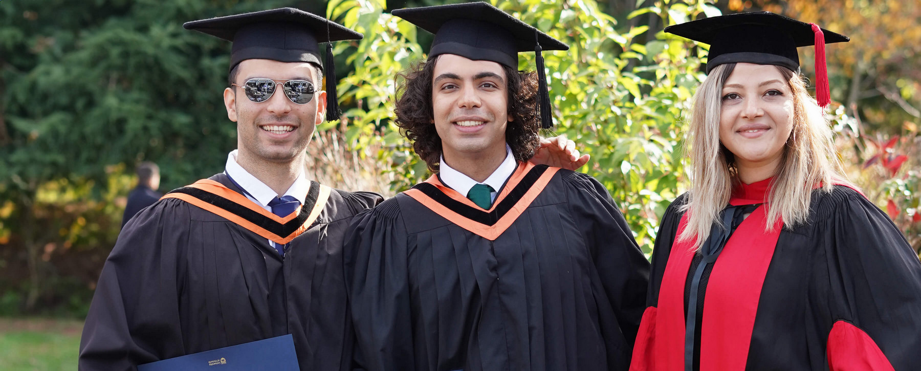 Three students wearing graduation caps and gowns standing outside and smiling..