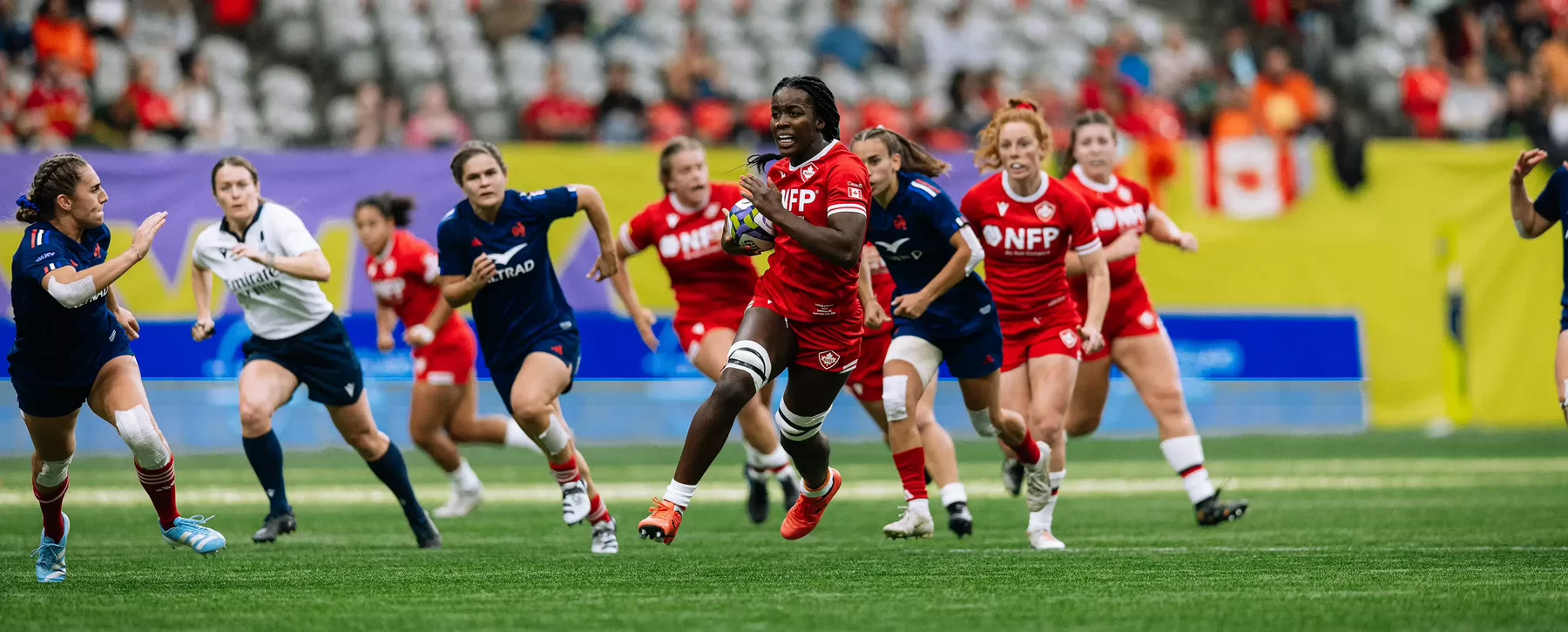 A Canadian women's rugby team player runs with the ball past a crowd of opponents. 