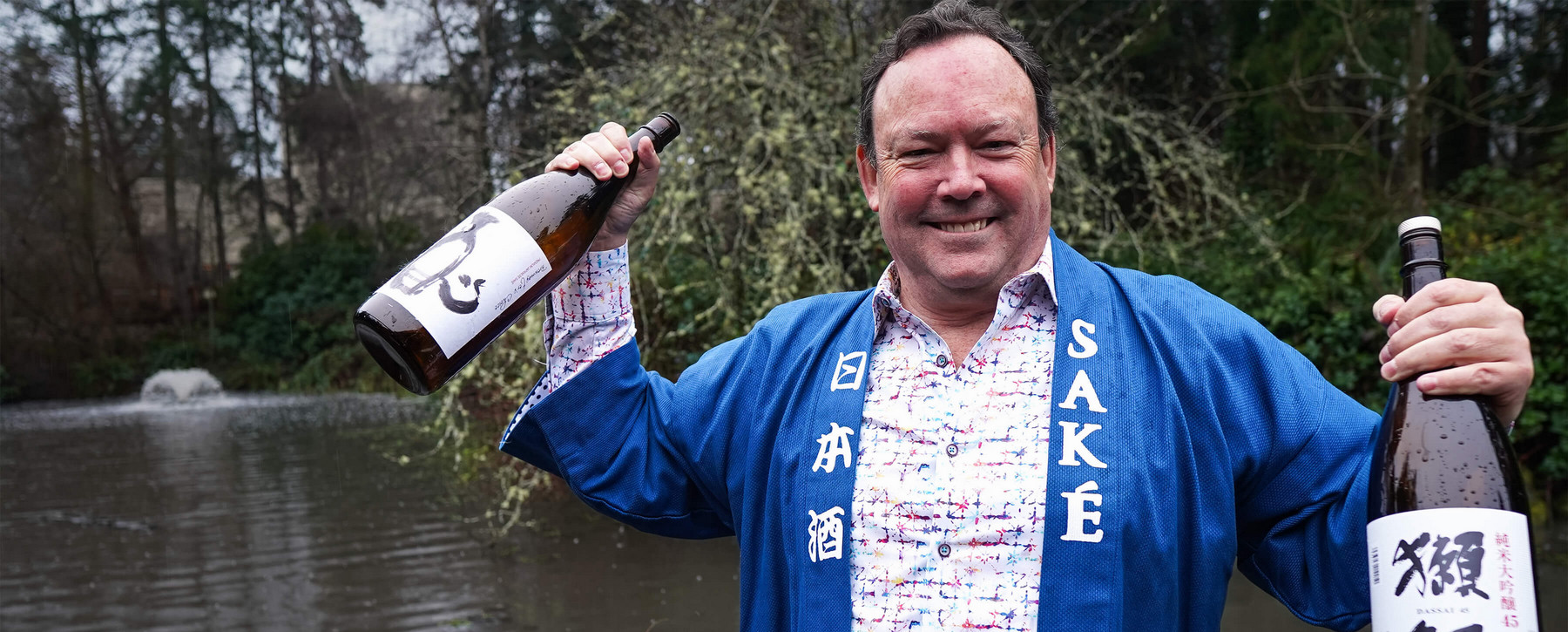Man wearing a Japanese robe that says Sake on the lapel, smiling and holding up a bottle of sake in each hand.