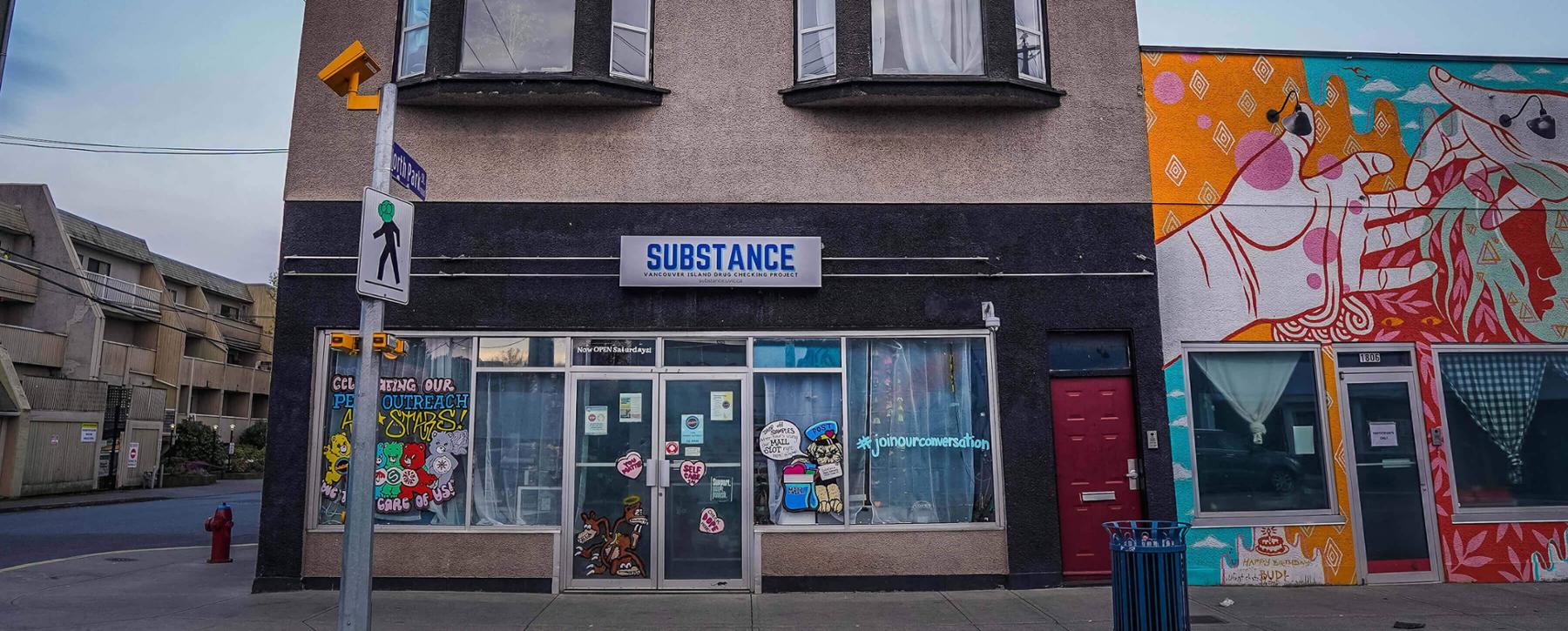 Storefront of a two storey grey stucco building with drawings of cartoon bears on the windows and a neighbourhing building decorated with mural art of two outreached hands touching. 
