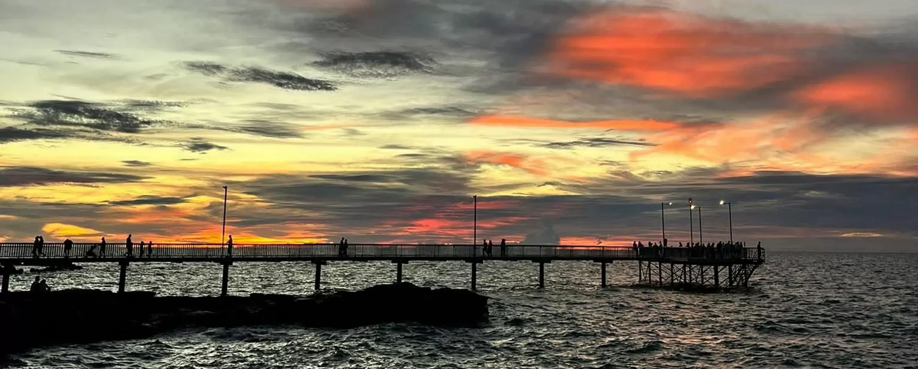 Silhouettes of people walking on a pier over the ocean as the sun sets.