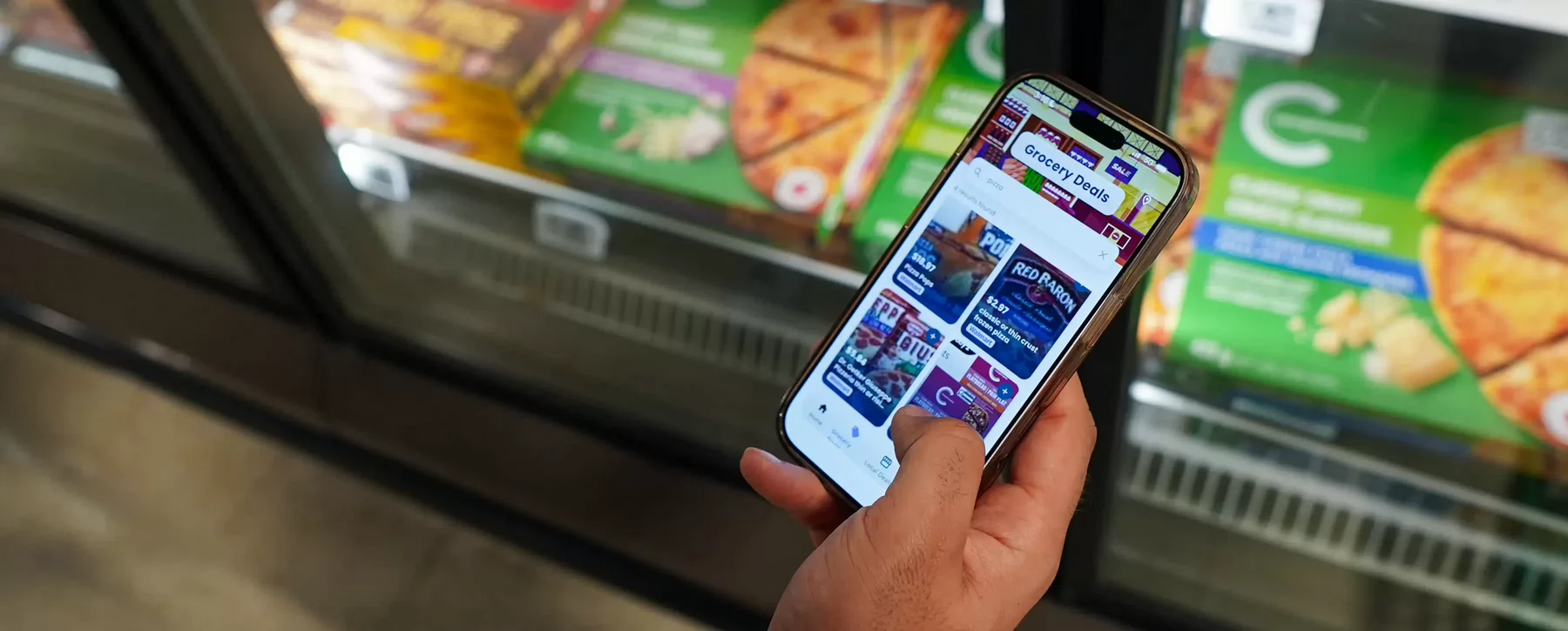 A hand holding a smartphone that says Grocery Deals on the screen in front of a grocery freezer window. 