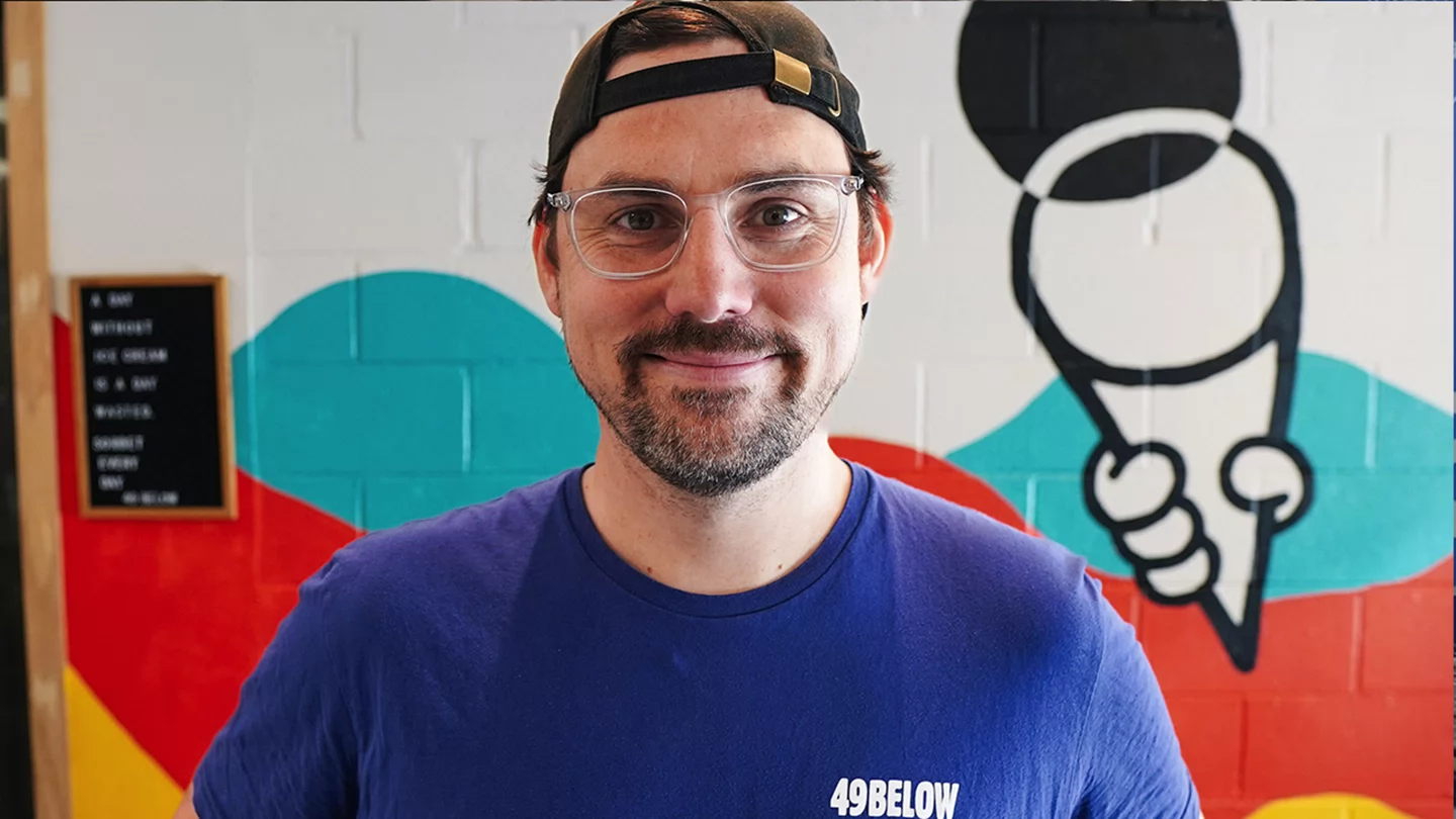 Man wearing glasses, backwards baseball and T-shirt that says 49 Below Ice Cream smiling while standing in front of colourful mural of an ice cream cone.  
