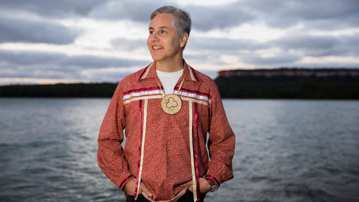 Man standing on the shore wearing decorative long sleeve shirt and lanyard.