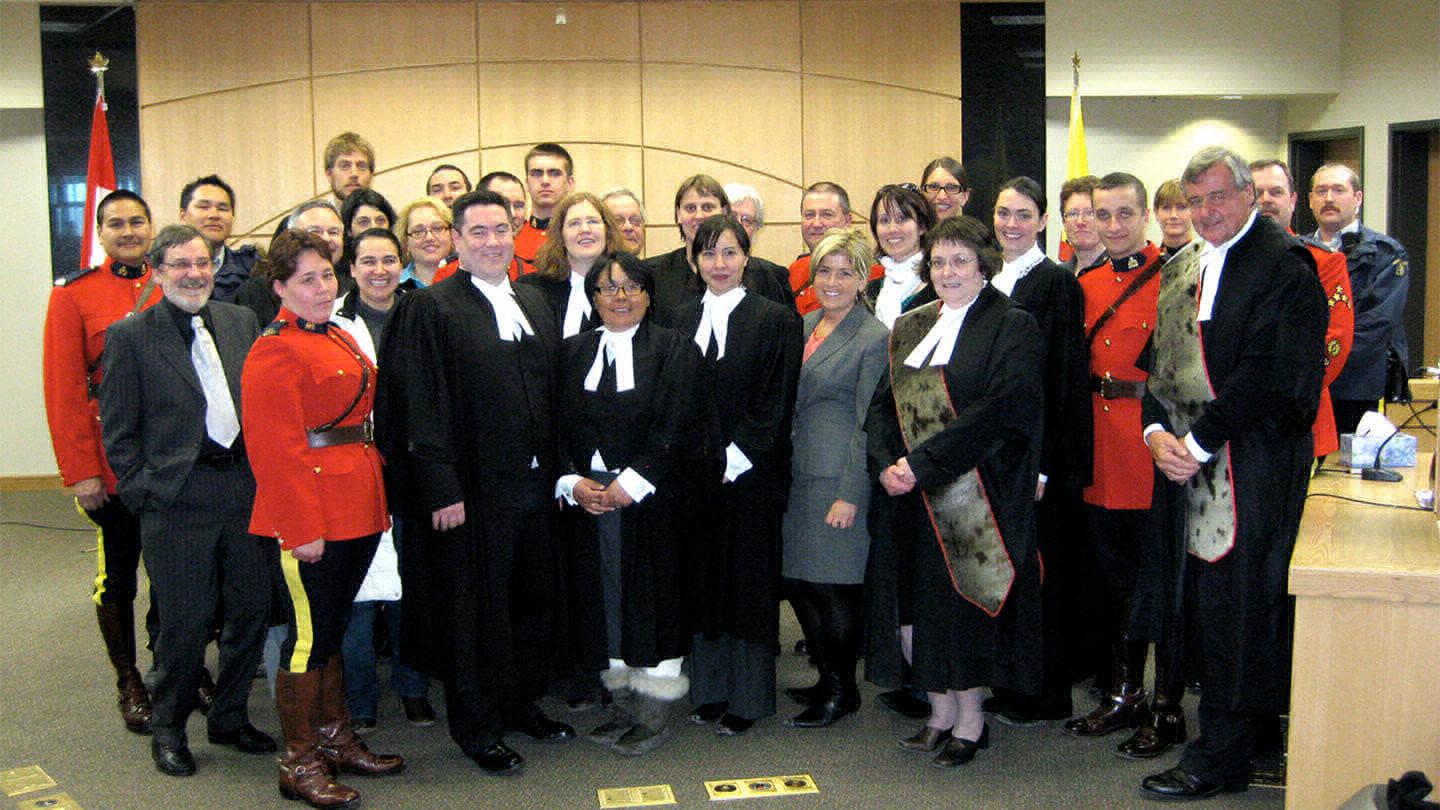 Group of people smiling and wearing lawyer robes standing with people in Canadian Mountie uniforms in a courtroom. 