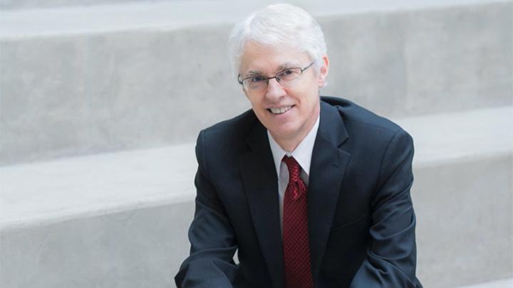 White haired man wearing glasses and suit smiling while sitting on concrete stairs.