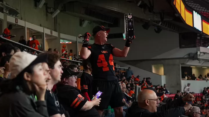A man wearing a BC Lions jersey bangs a drum in the stands of a BC Lions football game.