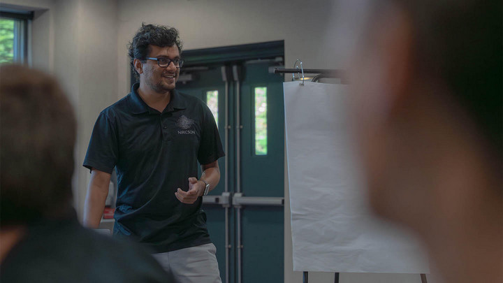 Man in black shirt wearing glasses beside white drawing board speaking to room of people.